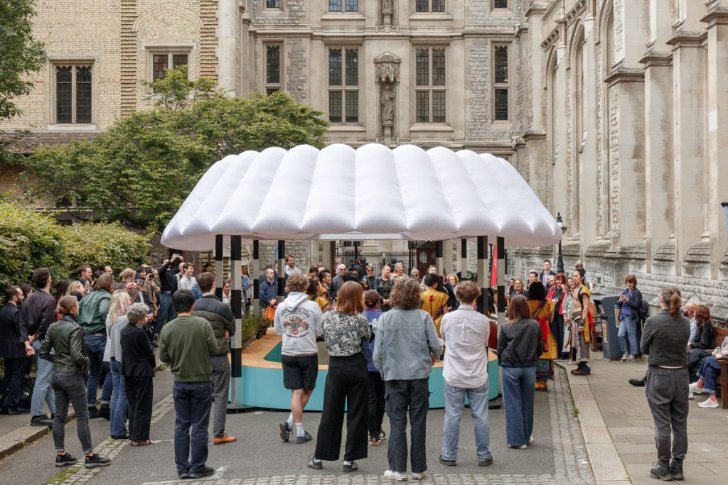 A crowd of people gather around a structure with an inflatable roof to watch a performance of drummers. In the background is a gothic library building