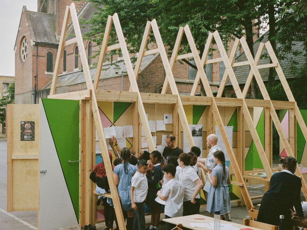 Children stand next to a wooden structure with drawings pinned on.